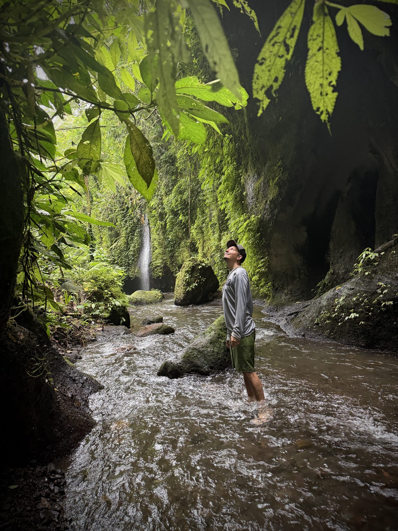 A person standing in a stream in a lush green forest with a waterfall behind